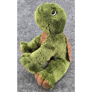 Puede incluir: Un juguete de peluche con forma de tortuga que presenta un pelaje verde oliva suave, un panel de pecho color canela liso y un caparazón marrón texturizado en la espalda. El peluche tiene ojos grandes y expresivos y patas acolchadas marrones.
