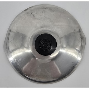 Könnte beinhalten: Ein runder, silberfarbener Topfdeckel aus Metall mit einer glatten, reflektierenden Oberfläche. Der Deckel verfügt über einen mittig platzierten, schwarzen, runden Knopf mit einem gestuften, gerillten Design. Die Metalloberfläche weist Kratzer auf.