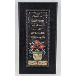 May include: Framed wall art with the text "Write it on your heart that the ones you love are life's most precious gifts." The image shows a floral arrangement in a pot with red and blue flowers, set within a black frame.