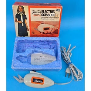 Könnte beinhalten: Vintage Scovill Dritz elektrische Schere in Originalverpackung. Die Schachtel zeigt ein Bild einer Frau und den Text "ELECTRIC SCISSORS". Die Schere ist weiß mit einem orangefarbenen Akzent und hat ein Kabel.