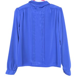 May include: A long-sleeved, cobalt blue blouse with a high, draped neckline. The front of the blouse features vertical pleats. The sleeves have buttoned cuffs. The blouse is hanging on a pink hanger against a white background.