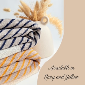 May include: Folded blankets in navy blue and yellow stripes. The blankets are beige and white. The text "Available in Navy and Yellow" is on the right. A beige vase with dried flowers is in the background. The blankets are folded.