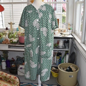 May include: A short-sleeved house dress in a deep forest green color featuring a repeating white paisley pattern. The garment has a full-length front button closure with white buttons, a rounded neckline, and two large patch pockets on the front.