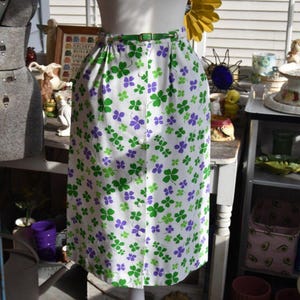 May include: A white skirt with a green belt and a pattern of green and purple shamrocks. The skirt has a button-up front and is knee-length. The skirt is made of a lightweight fabric.