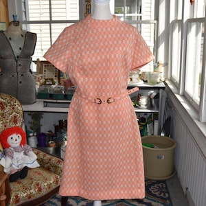 Puede incluir: Un vestido vintage color melocotón con un estampado geométrico a cuadros. El vestido presenta mangas cortas, cuello alto y un cinturón a juego con hebilla dorada. El vestido se exhibe en un maniquí.