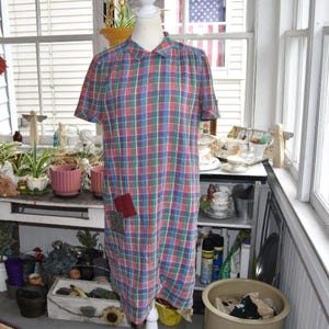 May include: A short-sleeved dress featuring a multi-colored plaid pattern in shades of blue, green, and red. The dress has a classic collar, a button-down front, and two small, contrasting fabric patches sewn onto the lower front section.