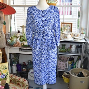 May include: A long-sleeved dress featuring a blue and white floral pattern, a round neckline, and a button-down front closure. The garment includes a peplum waist detail and a straight skirt that falls to a midi length, crafted from a lightweight fabric.
