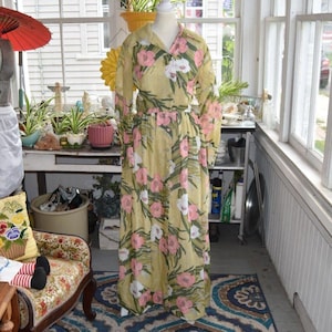 May include: A long-sleeved, floor-length dress in a pale yellow hue, featuring a repeating floral pattern of pink and white blossoms with green foliage. The garment has a collared neckline, a button-down front, and a cinched waistline.