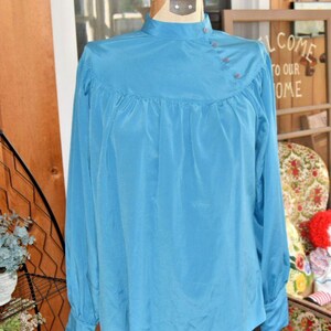 Pode incluir: Uma blusa azul-turquesa com mangas compridas e gola alta. A blusa apresenta um detalhe franzido no peito e três pequenos botões no ombro. As mangas têm punhos abotoados. A blusa é exibida em um manequim.