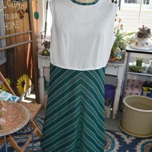 Puede incluir: Un vestido vintage sin mangas en un maniquí. La parte superior es blanca con un ribete verde alrededor del escote. La falda es verde con rayas de chevron blancas. El vestido se exhibe en una habitación con un letrero de madera que dice "Bienvenido a casa".
