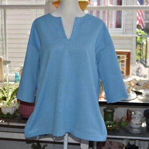 May include: A light blue top with a textured waffle-knit pattern, featuring a relaxed fit, three-quarter length sleeves, and a notched V-neckline. The garment has a straight hemline and a consistent grid-like weave throughout the fabric.
