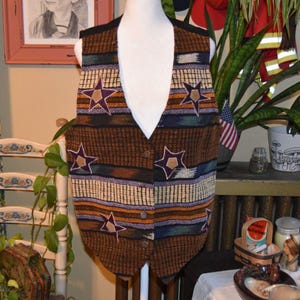 Peut inclure: Un gilet sans manches présentant un motif tissé de rayures horizontales dans des tons marron, beige, bleu et violet. Le vêtement est orné de plusieurs appliqués en forme d'étoile à cinq branches violettes et beiges, avec quatre boutons.