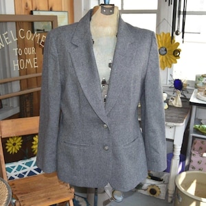 May include: A gray blazer with two buttons, displayed on a mannequin. The blazer has a notched lapel and two front pockets. The blazer is made of a textured fabric. The background includes a wooden chair and a sign that says "WELCOME TO OUR HOME."