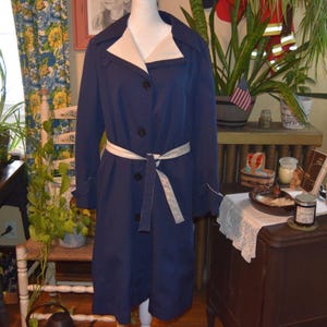 May include: A navy blue trench coat featuring a contrasting cream-colored wide collar and a matching cream-colored tie belt at the waist. The coat has long sleeves with cuff details, a button-down front closure, and a knee-length hemline.
