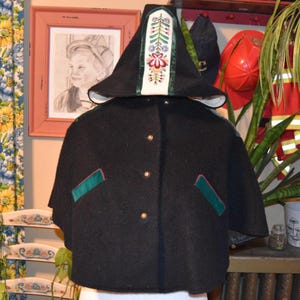 May include: A black wool cape and hat set. The cape has gold buttons and green trim. The hat features a white panel with colorful floral embroidery. The set is displayed on a mannequin, showcasing its design and details.