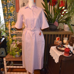 May include: A light purple dress with short sleeves and a matching belt. The dress has a collared neckline and a button-down front. A small tag is attached to the pocket. The dress is displayed on a mannequin.