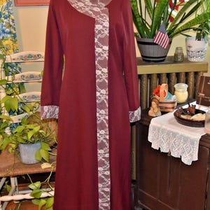 May include: A burgundy-colored dress with long sleeves and a round neckline. The dress features white lace trim along the front, neckline, and sleeve cuffs. The dress is displayed on a mannequin.