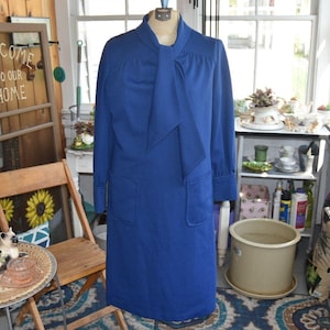 May include: A solid royal blue long-sleeve dress featuring a high neckline with an attached neck tie, long sleeves with cuffs, and two large patch pockets on the front. The dress has a straight silhouette and is displayed on a dress form.