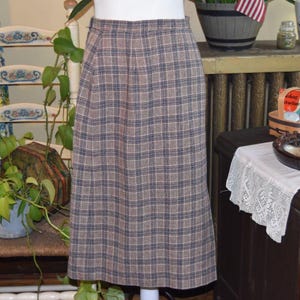 May include: A vintage knee-length skirt with a plaid pattern in brown, beige, and navy blue. The skirt is displayed on a mannequin. The skirt's design is a classic plaid.