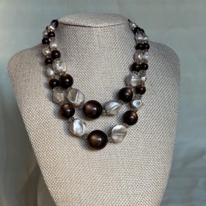 May include: A two-strand necklace with large, round, and irregular beads in shades of brown, cream, and clear. The necklace is displayed on a beige mannequin bust. The beads are strung together, creating a layered effect.
