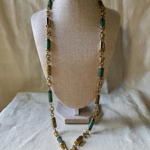 May include: A long necklace featuring a gold-tone chain with alternating cylindrical green stone beads and ornate gold-tone metal spacers. The design incorporates a repeating pattern of smooth green segments and textured metallic links throughout the length.