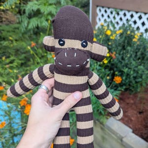 May include: A handmade sock monkey doll in brown and tan stripes. The monkey has button eyes and a stitched mouth. The doll is being held up in front of a background of green foliage and orange flowers.