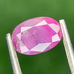 Könnte beinhalten: Ein facettierter, ovalförmiger Rubin-Edelstein, der von einer silbernen Pinzette gehalten wird. Der Rubin zeigt einen leuchtenden rosa Farbton mit weißen Facetten, vor einem verschwommenen grünen Hintergrund. Die Oberfläche des Edelsteins weist kleine Einschlüsse auf.