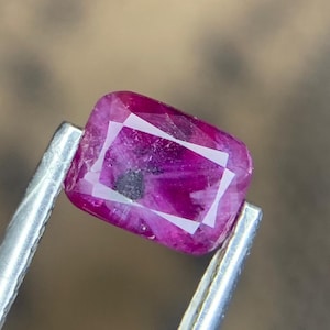 Puede incluir: Un vibrante rubí rectangular, sujeto con pinzas plateadas. El rubí muestra un tono magenta intenso con inclusiones internas y facetas. La gema es el punto focal, mostrando su belleza natural y potencial para joyería.