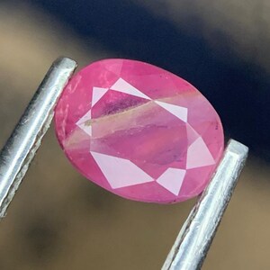Könnte beinhalten: Ein facettierter, ovalförmiger Rubin-Edelstein, gehalten von einer silbernen Pinzette. Der Rubin zeigt einen leuchtenden rosaroten Farbton mit inneren Einschlüssen. Der Edelstein ist so geschliffen, dass er das Licht optimal reflektiert.