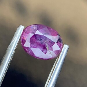 Puede incluir: Una gema facetada de color rojo púrpura, de forma ovalada, sujeta con pinzas plateadas. La gema tiene una superficie pulida con inclusiones internas visibles. Las pinzas están enfocadas, resaltando la claridad y el color de la gema.