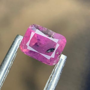Könnte beinhalten: Ein facettierter, rechteckiger rosa Edelstein mit weißem Innenrand wird von einer silbernen Pinzette gehalten. Der Stein hat eine leicht unregelmäßige Form und einen dunklen Einschluss. Der Hintergrund ist verschwommen, mit Braun- und Dunkelblautönen.