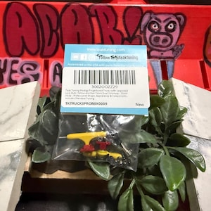 Puede incluir: Un paquete de ejes para fingerboard con tuercas de seguridad mejoradas, ejes amarillos y rojos y una cubierta antipolvo. El paquete está etiquetado con información del producto frente a un fondo rojo con graffiti.