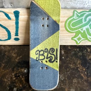 Puede incluir: Una pequeña tabla de fingerboard con un diseño geométrico amarillo y gris. La tabla tiene el texto "B2S!" en negro. Tiene cuatro pequeños agujeros para los ejes y mide unos 10 cm de largo.