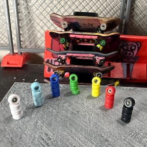 Puede incluir: Una colección de monopatines en miniatura apilados unos sobre otros, con una variedad de ruedas de colores dispuestas delante. Los monopatines tienen un aspecto desgastado y las ruedas son blancas, azul claro, azul, verde, amarillo, rojo y negro.