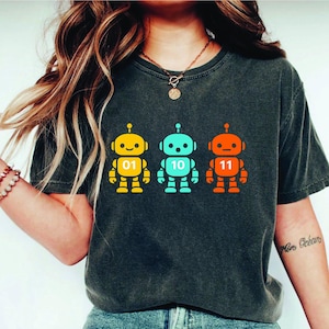 以下が含まれることがあります： 3つのカラフルなロボットデザインが施された濃いグレーのTシャツ。各ロボットは黄色、ターコイズ、オレンジ色で、胸には01、10、11の数字が表示されています。ロボットにはアンテナがあり、立っています。