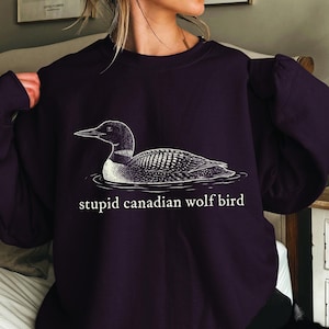 Pode incluir: Camisola roxa escura com uma ilustração detalhada a preto e branco de um mergulhão na água. O texto "stupid canadian wolf bird" está impresso abaixo. A camisola tem mangas compridas.