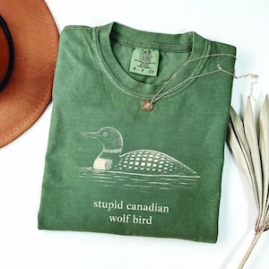 Pode incluir: Camiseta verde com um gráfico de um mergulhão na água e o texto "stupid canadian wolf bird". A camisa tem gola redonda e uma etiqueta pequena. Um colar de ouro com um pingente e um chapéu marrom também estão na imagem.