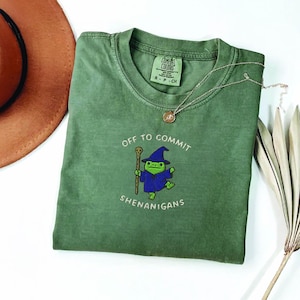 May include: Green t-shirt featuring the text "Off to Commit Shenanigans" and a cartoon frog in a wizard outfit. A gold necklace and a brown hat are also visible.