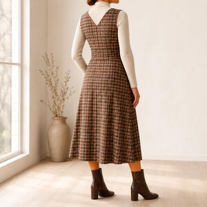 May include: A sleeveless midi-length dress featuring a brown, orange, and black plaid pattern with a V-neckline and a defined waistline. The dress is layered over a long-sleeved cream-colored turtleneck top and paired with dark brown leather ankle boots.