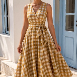 K&ouml;nnte beinhalten: Ein &auml;rmelloses Midikleid mit einem senfgelben und wei&szlig;en Vichy-Karomuster. Das Kleid hat einen V-Ausschnitt, ein eng anliegendes Oberteil und ein Taillenband, das vorne zu einer Schleife gebunden ist. Der Rock ist weit und plissiert, wadenlang.