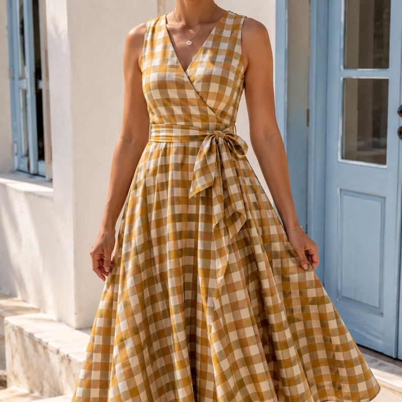 K&ouml;nnte beinhalten: Ein &auml;rmelloses Midikleid mit einem senfgelben und wei&szlig;en Vichy-Karomuster. Das Kleid hat einen V-Ausschnitt, ein eng anliegendes Oberteil und ein Taillenband, das vorne zu einer Schleife gebunden ist. Der Rock ist weit und plissiert, wadenlang.