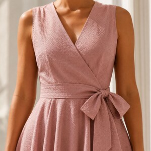 K&ouml;nnte beinhalten: Ein &auml;rmelloses, altrosa Kleid mit V-Ausschnitt und Wickeloberteil. Das Kleidungsst&uuml;ck verf&uuml;gt &uuml;ber ein passendes Stoffband in der Taille, das seitlich zu einer Schleife gebunden ist, und das Material weist eine subtile, texturierte Oberfl&auml;che auf.