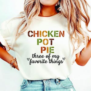 May include: Cream-colored t-shirt with the words "CHICKEN POT PIE three of my favorite things" in brown, green, orange, and black lettering. The shirt has a relaxed fit and is made of a soft material.