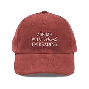 Puede incluir: Gorra de béisbol de pana rojo ladrillo con el texto bordado "ASK ME WHAT Book I'M READING" en blanco. La gorra tiene una visera curva y una correa ajustable.