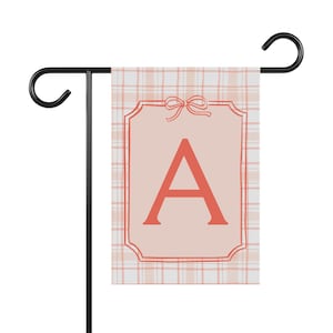 Könnte beinhalten: Gartenflagge mit einem korallenfarbenen Buchstaben "A" auf hellrosa Hintergrund. Die Flagge hat ein Karomuster und ist von einem dekorativen Rahmen und einer Schleife umgeben. Sie ist auf einem schwarzen Metallständer montiert.