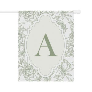 K&ouml;nnte beinhalten: Eine rechteckige Gartenflagge mit hellgrauem Hintergrund und einem botanischen Blumenmuster in Salbeigr&uuml;n. In der Mitte befindet sich ein dekorativer, geschwungener Rahmen, der einen gro&szlig;en, fetten Buchstaben A in Salbeigr&uuml;n umschlie&szlig;t.