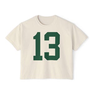 May include: Cream-colored t-shirt with the number 13 in a bold, dark green font. The shirt has a classic crew neck and short sleeves. The number is centered on the front of the shirt.