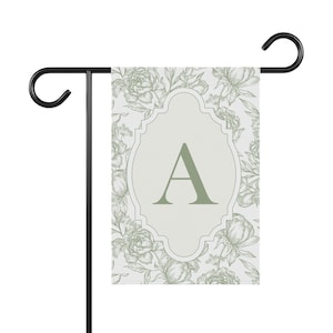May include: A rectangular garden flag featuring a light gray background with a sage green floral line art pattern. In the center, a decorative scalloped frame encloses a large, sage green capital letter A. The flag hangs from a black metal stand.
