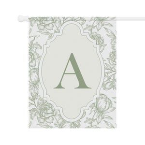 K&ouml;nnte beinhalten: Eine rechteckige Gartenflagge mit einem salbeigr&uuml;nen Blumenmuster als Strichzeichnung auf wei&szlig;em Grund. Ein zentraler, dekorativer Rahmen mit gewelltem Rand enth&auml;lt einen gro&szlig;en, fetten, salbeigr&uuml;nen Gro&szlig;buchstaben A. Die Flagge h&auml;ngt an einer wei&szlig;en Stange.