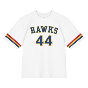 May include: White t-shirt with the word "HAWKS" in blue and gold lettering and the number "44" in blue and gold. The sleeves have red, yellow, and blue stripes. This shirt is a sports-themed apparel item.